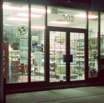 A pharmacy is seen through a glass storefront at night, brightly lit from inside. The doors and windows are transparent, revealing shelves stocked with various products. Signs are displayed on the glass, and there's a Coca-Cola vending machine visible. The interior lights highlight the organized arrangement of goods on the shelves.